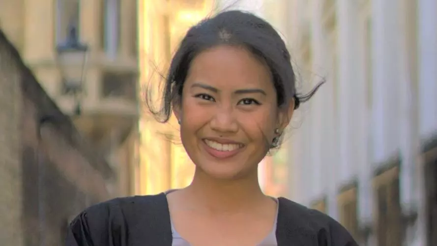 A smiling Marianne stands in a narrow street wearing a black graduation gown.