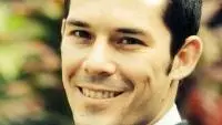 Matthew has short dark hair and smiling at the camera. He's wearing a smart checked white shirt with tie and jacket