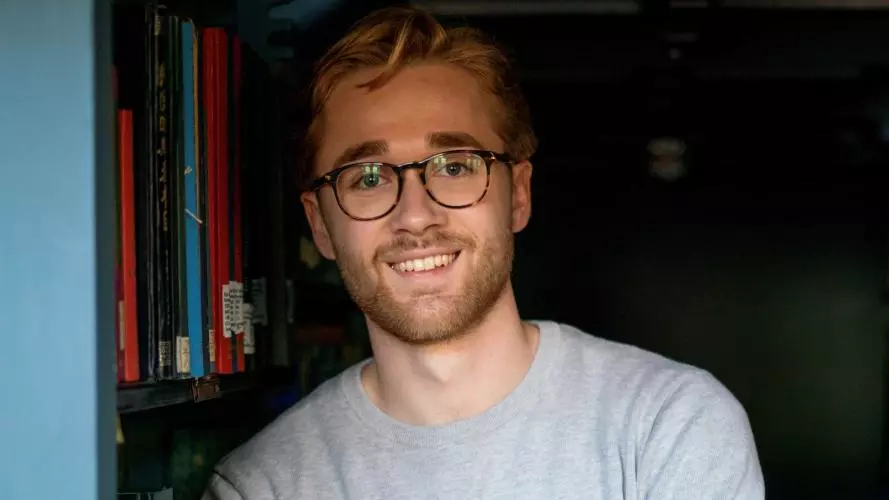 Robert Hill has short fair hair and is wearing round tortoiseshell glasses and a grey jumper. He is leaning against a bookshelf.