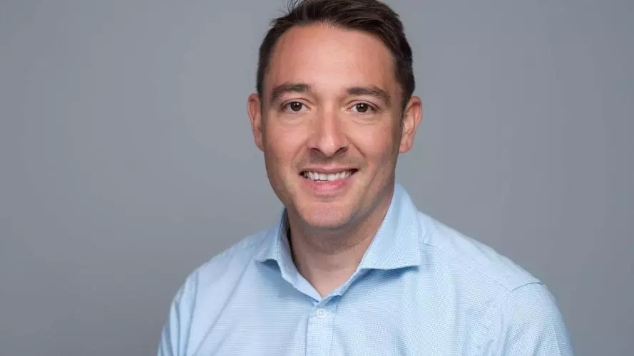 Man with dark brown hair and brown eyes, wearing a blue shirt and smiling