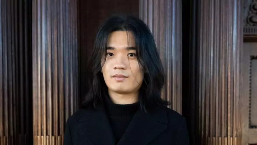 Xinghao li is wearing a black coat, with shoulder length hair looking at the directly at the camera.