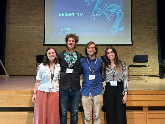 A group of four students standing with their arms around each other, in front of a raised stage and display screen that says 'SERSPI 2024'.