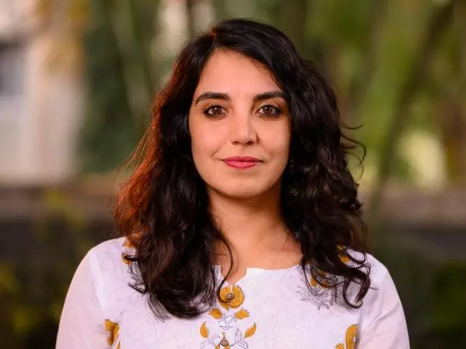 Wearing a pink top with mustard flower patterns and shoulder-length brown wavy hair, Anita smiles gently at the camera.