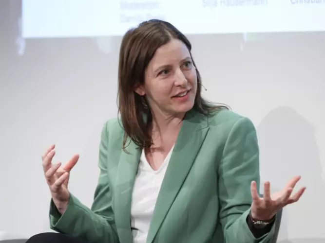 As Silja Häusermann sits on a stage wearing a green suit, she appears to be in a discussion and expressing herself with her hands. 