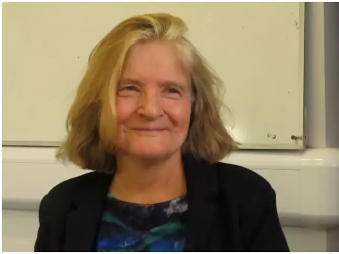 Professor Gardner has short blonde hair. She is wearing a dark blouse with a dark blue and green abstract pattern.