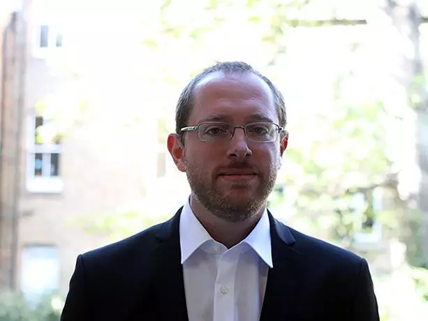 Man with light brown hair and gold rimmed glasses, wearing a white shirt and dark suit