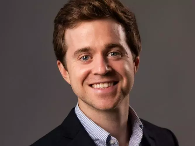 Zach Parolin has short brown hair. He is wearing a light blue shirt and a dark blazer, and smiling broadly. 