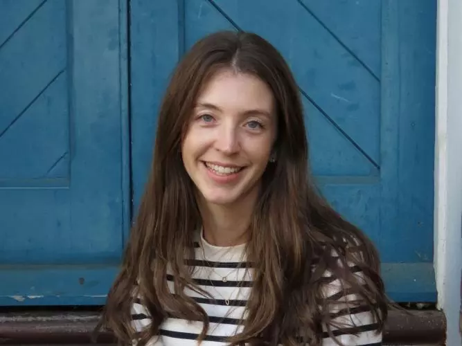 Bridget Steele has long brown hair and is wearing a black and white striped shirt. She is sitting on some outdoor steps in front of a blue door and smiling.