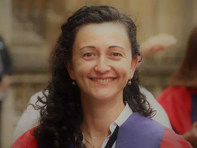 Elona  has long dark curly hair and is smiling at the camera. She's wearing a blouse under a purple and red jacket