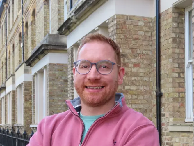 Dr Fanner has short fair hair and is wearing glasses. He is standing with his arms crossed, wearing a pink sweater. 