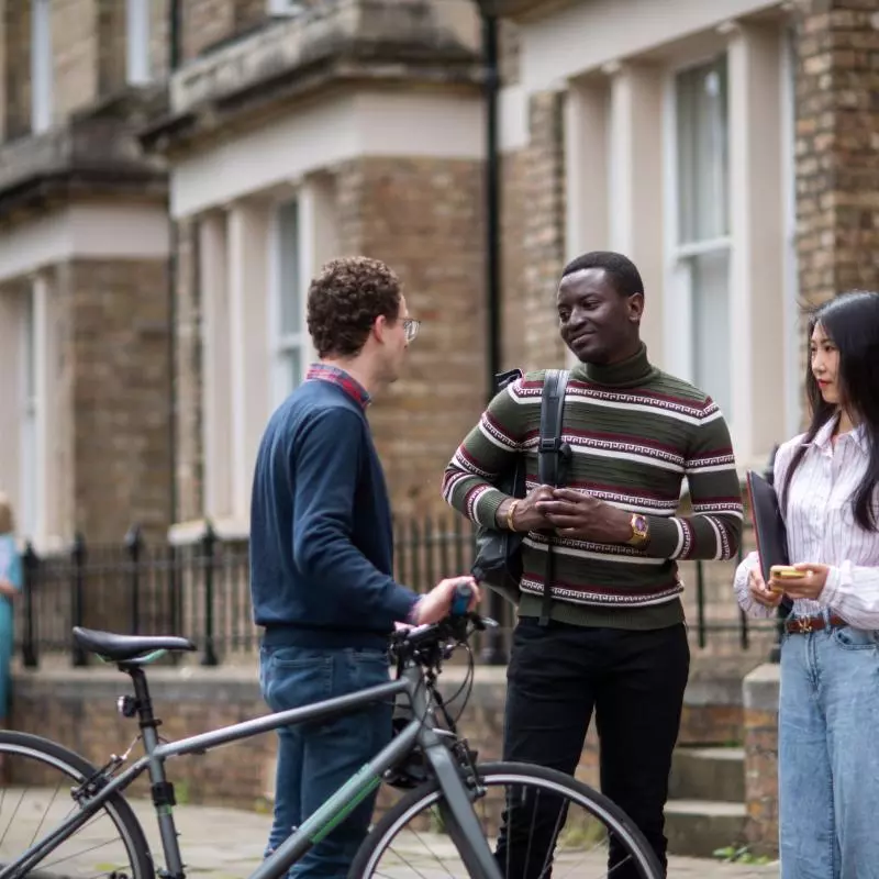 Three students standing outside department buildings. A male student on the left is holding a bicycle, facing the two other students. 
