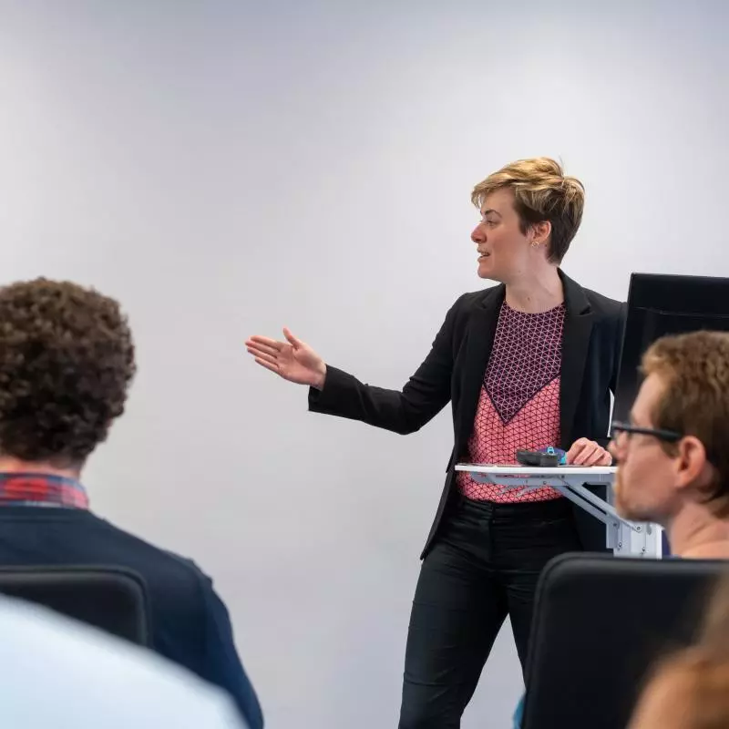 A lecturer standing in front of a class, looking and gesturing to the side
