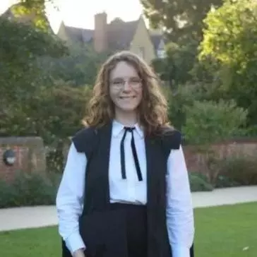 Nicola Tomlin wearing full academic dress and is standing in a college garden.  Nicola has long brown curly hair and wearing glasses