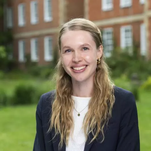 Sophia has long blonde hair. She is wearing a white shirt and navy blazer. She is sat outside in a garden with a red-brick building behind her.