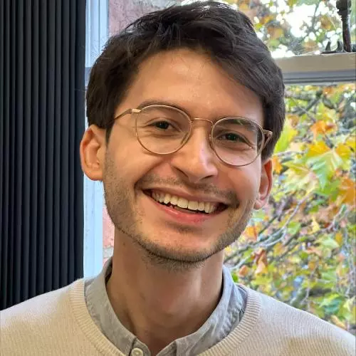 David has brown hair, gold rimmed glasses and is smiling at the camera. He is wearing a cream jumper and grey shirt.