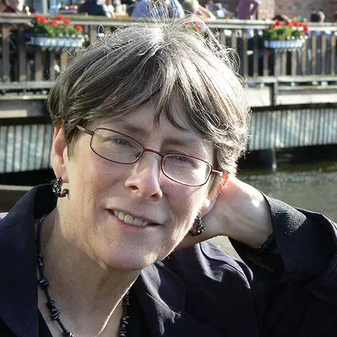 A woman with dark grey hair and metal rimmed glasses sits on a bench with a canal in the background.