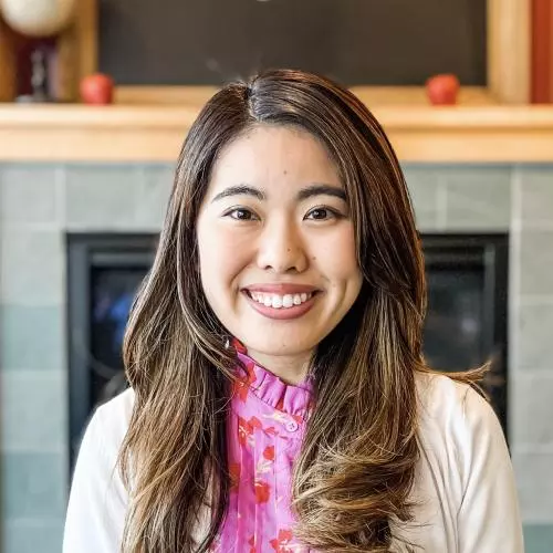 Hinako has long dark brown hair and is smiling at the camera. She's wearing a pink patterned blouse with a white cardigan. 