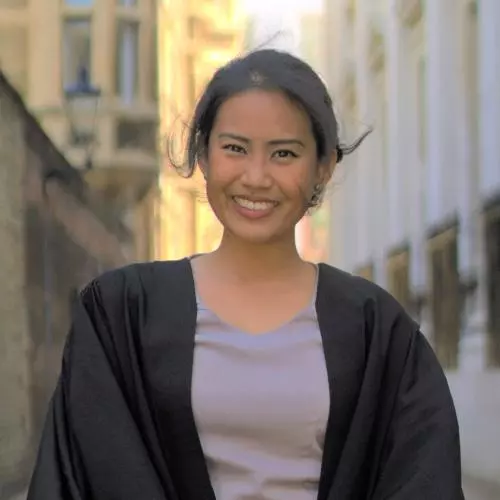 Marianne is standing in a narrow street, wearing a black graduation gown with her hair tied back and smiling.
