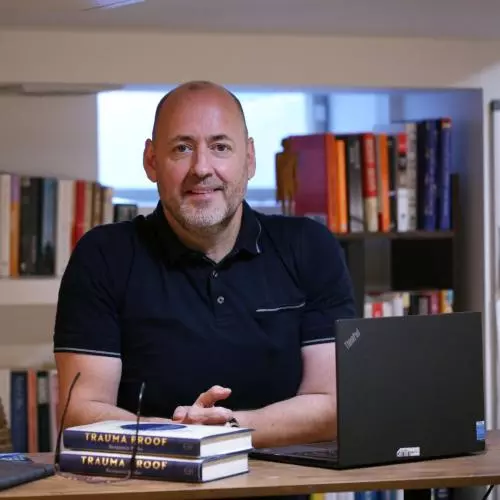 A smiling man seated at a desk in front of bookshelves, wearing a dark short-sleeved polo shirt.