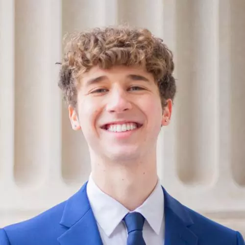 August is smiling and wears a blue suit and tie