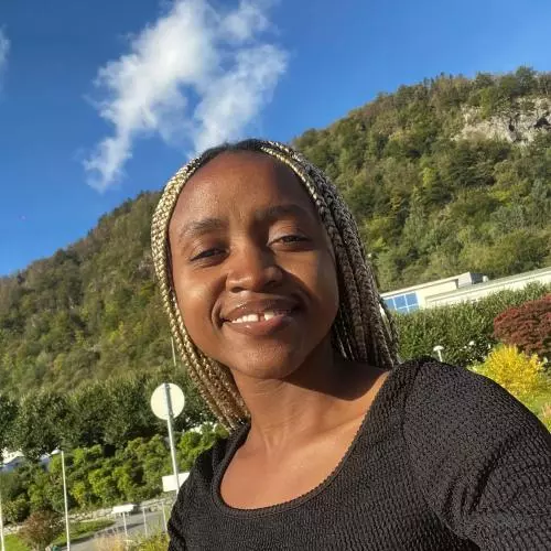 Diana Mwala is smiling in front of a blue sky. Her hair is braided and there is a tree-lined hill in the background. 