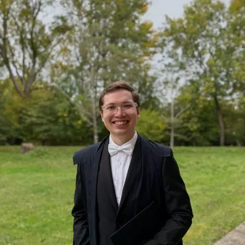 George Rappa has short brown hair and glasses. He's wearing an academic gown, with dark suit and white tie, and is standing in a college garden