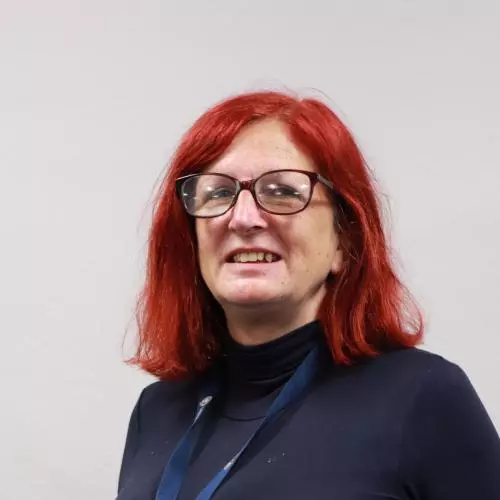 Woman with red hair and red rimmed glasses, who is smiling and wearing a dark top