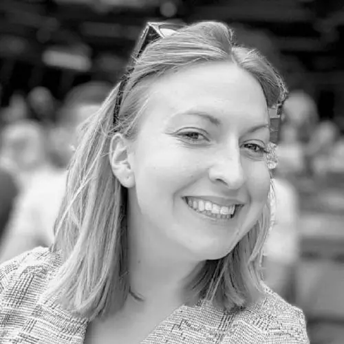 kathryn has shoulder-length blonde hair that is held back from her face by glasses on her head. She's smiling at the camera and is wearing a checked, woven jacket 