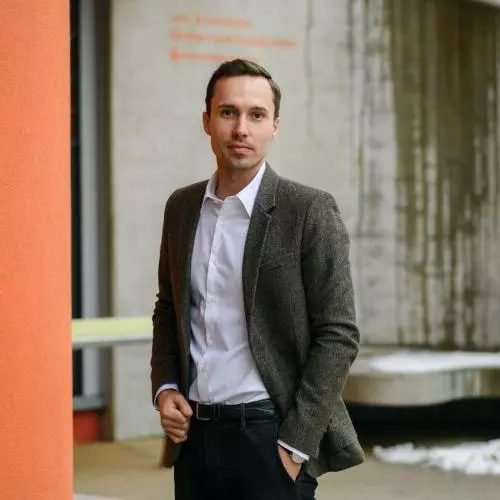 Lukas Lehner wears a white shirt and dark jacket, he stands on an angle next to an orange wall.