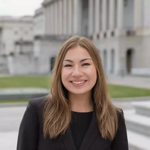 Miya has shoulder-length light brown hair and wearing a dark blazer. She is standing in front of a set of steps