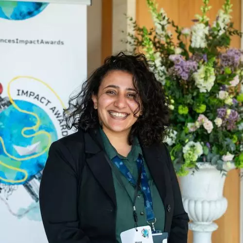 Mona Ibrahim has long curly dark hair. She is smiling broadly, standing in front of a bouquet of flowers and an 'Impact Awards' banner