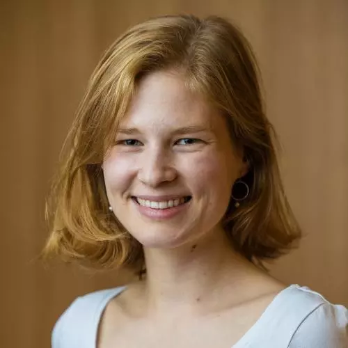 Sara Hueber has bobbed ginger hair and is wearing a white top. She is in front of a brown background, smiling. Credit: Tom Weller Photography, 2025