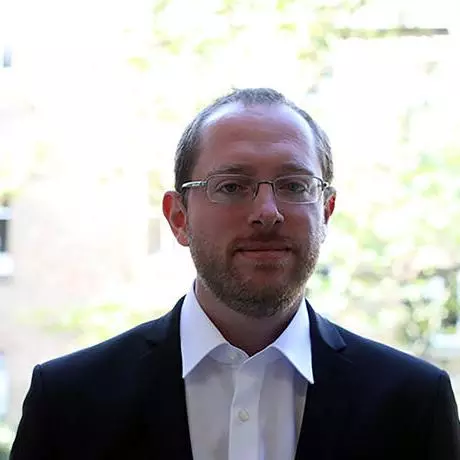 Man with light brown hair and gold rimmed glasses, wearing a white shirt and dark suit