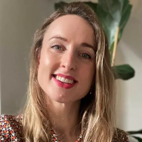 Adrienne has long blonde hair and is smiling at the camera. She's wearing a top with small white, orange and brown patterns.