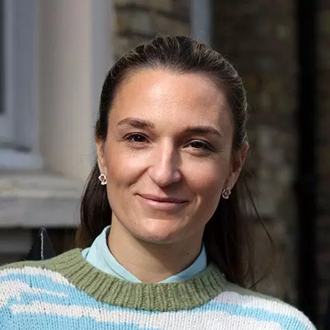 Alice has long dark hair swept back from her face. She is wearing a green and pale blue jumper