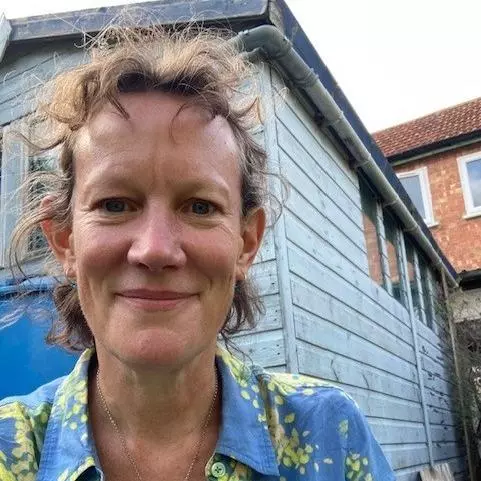 Amanda has curly brown hair which is partially tied back. She is wearing a patterned green and blue blouse, and sat outside in front of a small wooden structure.