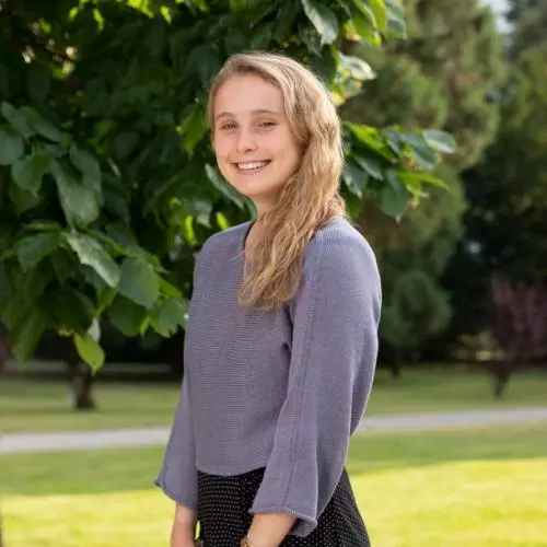 Charlotte Evans is wearing a purple top , standing sideways at the camera, with shoulder length blonde hair smiling at the camera.