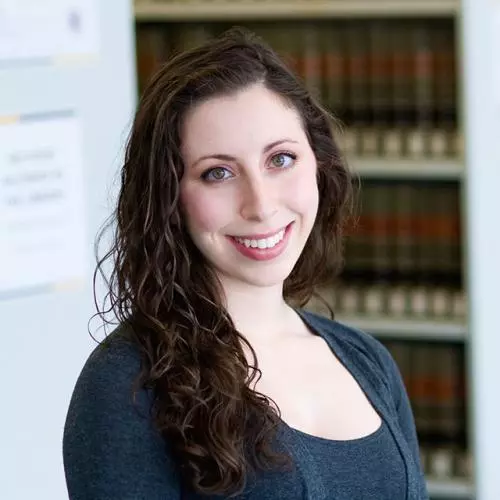 Alex has long dark curly hair. She is standing in front of some bookshelves, wearing a navy blue top and cardigan