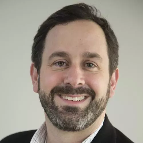 David has short dark hair and a beard, he is smiling at the camera, and wearing a white checked shirt with a dark jacket. 