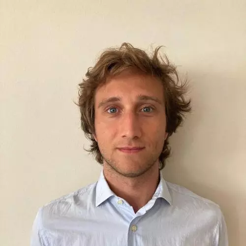 Enrico is standing in front of a wall looking at the camera. He has fair hair and is wearing a light shirt 