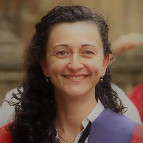 Elona  has long dark curly hair and is smiling at the camera. She's wearing a blouse under a purple and red jacket
