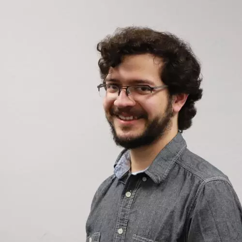 Man with curly dark hair and beard and wearing dark rimmed glasses and grey shirt