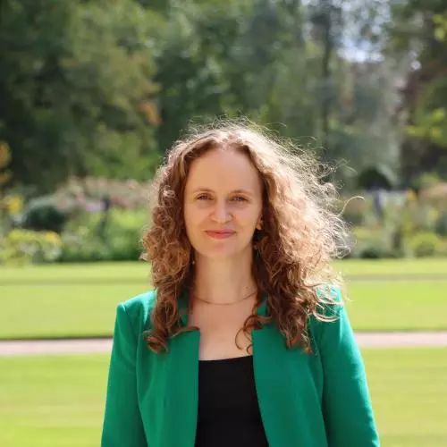 Jessica Lorimer has long curly brown hair. She is wearing a black dress and bright green blazer. She is standing outside in a large garden. 