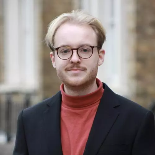 Hendrik Schäfer has blonde hair and wears glasses. He is standing in front of the DSPI building in a red polo-neck top and blazer.
