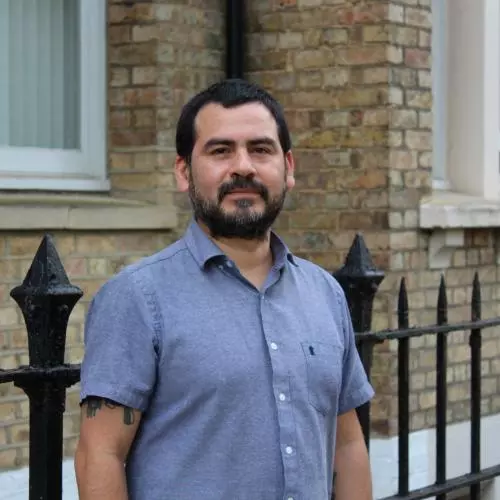 Dr Munoz has short black hair and black facial hair. He is wearing a short-sleeved blue shirt and standing outside a brick building. 