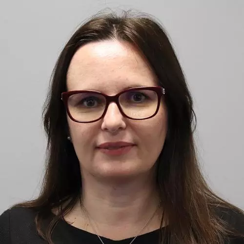 Woman with long brown hair, dark rimmed glasses and wearing a black top with necklace
