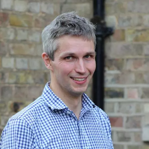 Jonathan is smiling at the camera. He has short grey hair and is wearing a blue and white checked shirt. 