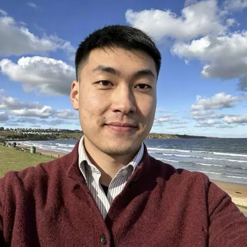Man with short dark hair, wearing a jumper and standing near the sea