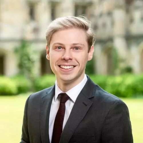 Madison Little has short blonde hair. He is wearing a white shirt, dark tie and blazer. He is standing in front of some grass in a courtyard.