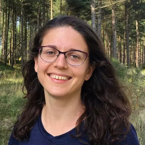 Yulia Shenderovich has long dark wavy hair, glasses and wearing a dark top. There is a background of trees behind her